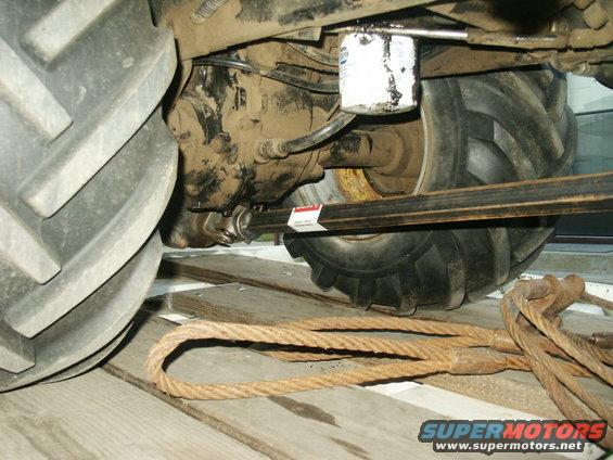 Undercarriage of a tractor showing the drive shaft, oil filter, and large tire. Ropes and wooden planks are in the foreground