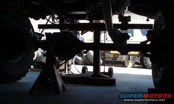 Underside view of a vehicle's rear axle and differential, supported by jack stands. Another vehicle is partially visible in the background