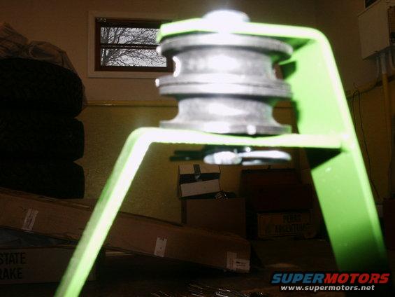 Green metal bracket holding a black pulley wheel, with stacked tires and boxes in the background
