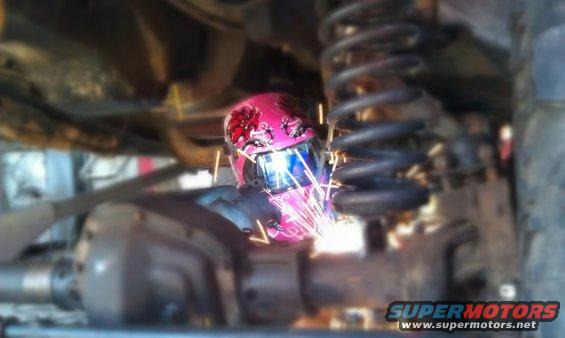 Welder with pink helmet sparks flying, working on vehicle suspension part