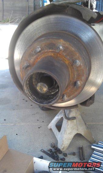 Close-up of a rusty brake rotor and hub assembly supported by a jack stand. Loose bolts are scattered on the ground