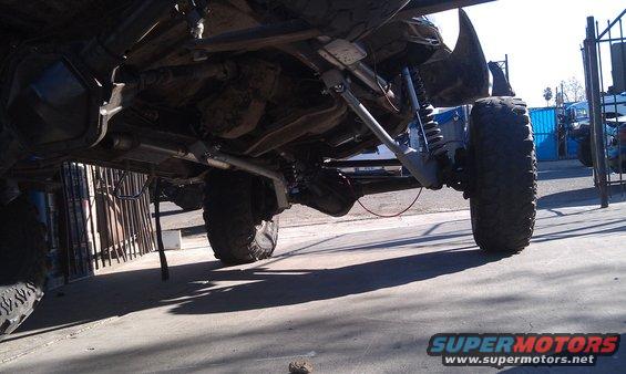 Underside view of a lifted off-road vehicle's suspension and drivetrain. Another vehicle is visible in the background