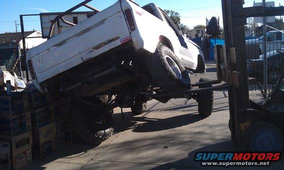 White pickup truck tilted on a forklift, with stacks of boxes underneath the rear