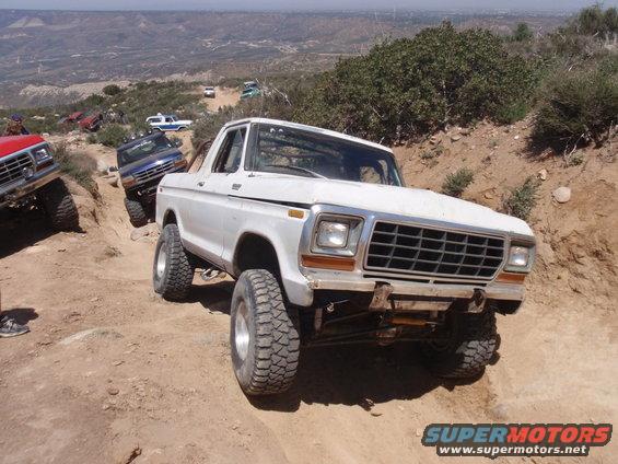 White 1978 Ford Bronco climbing a dirt hill with other off-road vehicles behind it