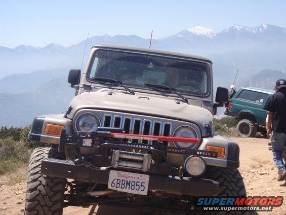 Gray Jeep Wrangler with winch, red Hi-Lift jack, and California plate 6BBM855. Green SUV in background