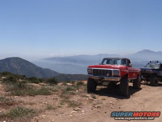 Red 1978 Ford F-150 pickup truck and a dark SUV parked on a dirt road with mountains in the background