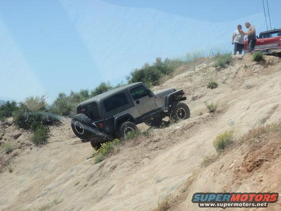 Gray Jeep Wrangler TJ climbing a dirt hill, two guys watching, red truck behind them