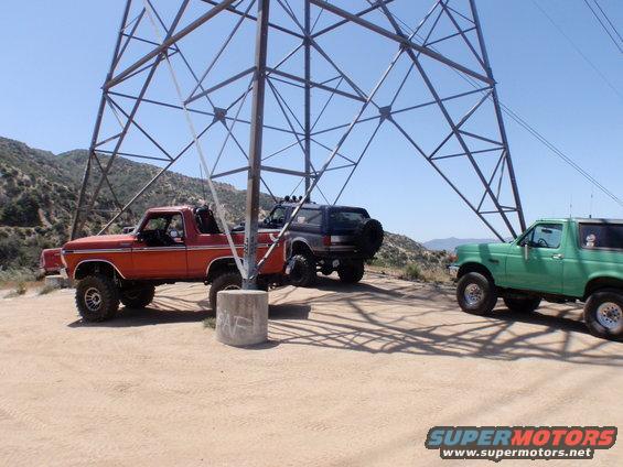 Three Ford Broncos parked under a power line tower. Orange Bronco in foreground, dark blue Bronco behind it, green Bronco to the right