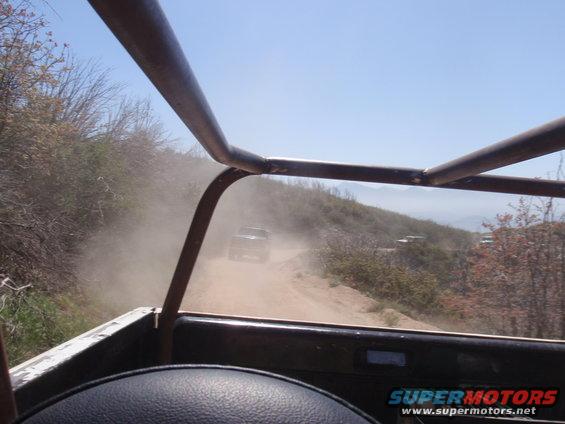 View from inside a vehicle looking out at a dusty off-road trail with other vehicles ahead