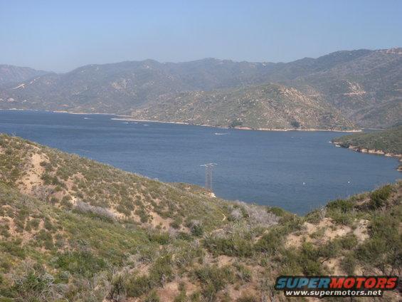 View of a large lake surrounded by hills and mountains under a clear blue sky. A power line tower is visible on the left
