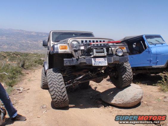 Gold Jeep Wrangler with large tires on a rock, red winch cable, blue Jeep in background