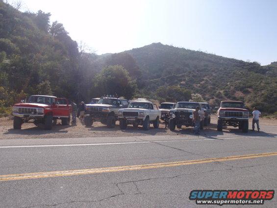 Several lifted Ford trucks and SUVs parked on a dirt shoulder, with a mountain in the background