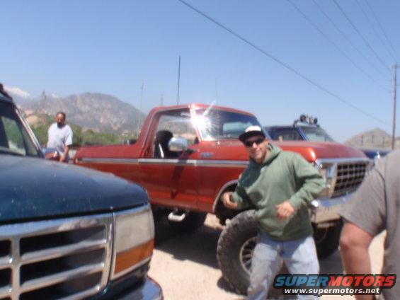 Red 1978 Ford Bronco parked next to a blue Ford F-Series truck. A man in a green hoodie stands by the Bronco