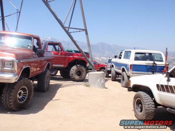 Several lifted trucks and SUVs, including a 1978 Ford Bronco, parked on a dirt hill with mountains in the background