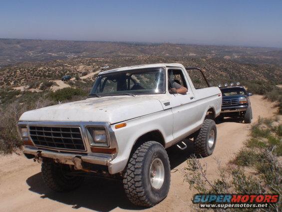 White 1978 Ford Bronco driving on a dirt road with another vehicle behind it