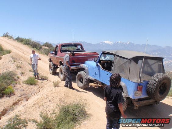Red 1978 Ford Bronco towing a blue Jeep on a dirt road with mountains in the background