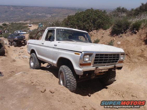 White 1978 Ford Bronco climbing a dirt hill with other vehicles behind it