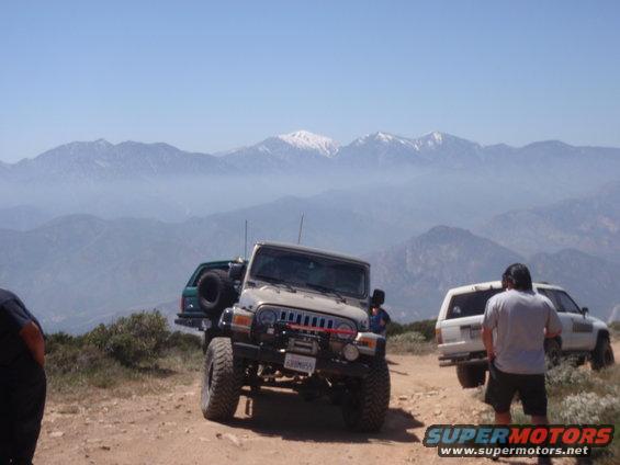 Tan Jeep Wrangler with winch and spare tire on rear, parked on dirt road with mountains in background