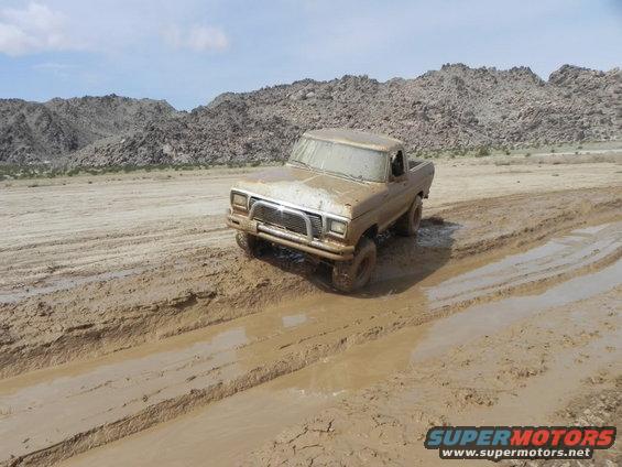 Mud-covered 1970s Ford F-Series truck driving through a muddy track in a desert landscape