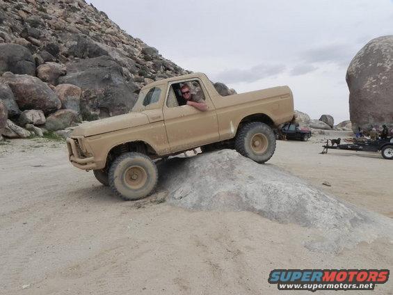 Tan 1978 Ford Bronco climbing a rock in a desert landscape. Driver visible