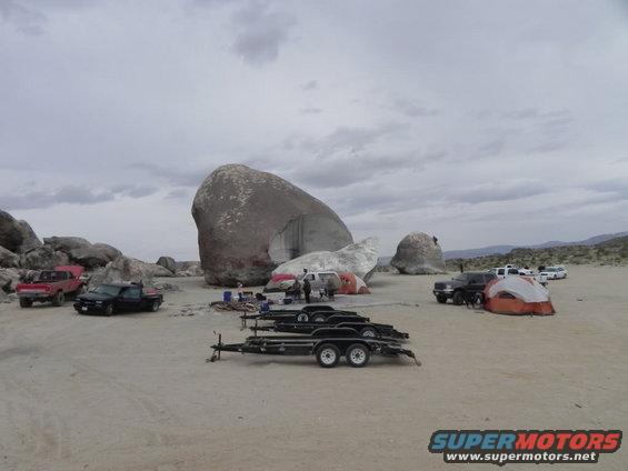 Desert scene with multiple trucks, trailers, and tents near large rock formations