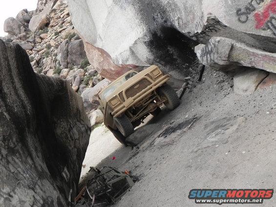Tan off-road vehicle climbing a steep, rocky incline next to large boulders. Another vehicle is visible below