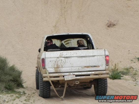 Rear view of a white 1978 Ford F-150 pickup truck driving on a dirt road, two people inside