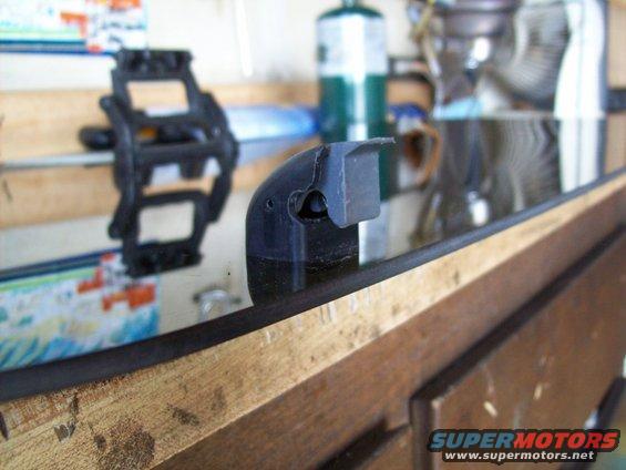 Black plastic part with a curved edge and a rectangular protrusion, resting on a wooden surface. A black pedal-like object is visible in the background
