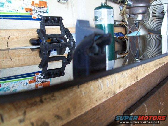 Black plastic clamp holding a metal rod on a wooden surface, with a propane tank and fan in the background