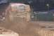 1978 Ford Bronco - A modified truck jumps through a mud pit, spraying dirt and water, with a tractor visible behind a green barrier.