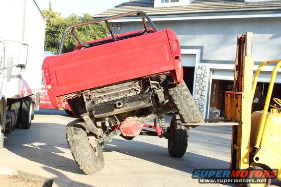 Red pickup truck tilted on forklift forks, rear view. Another truck is visible to the left