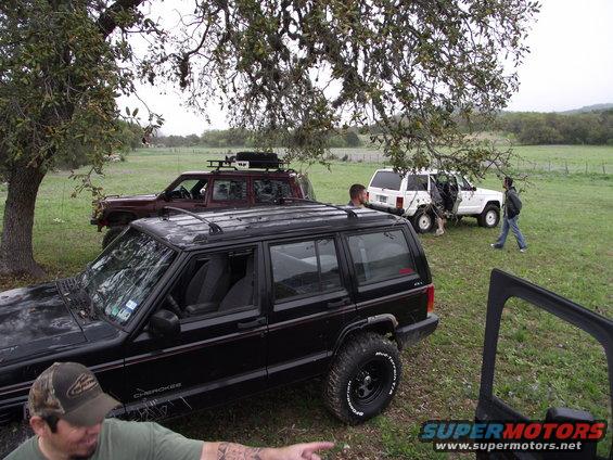 Black Jeep Cherokee XJ with off-road tires, parked in a grassy field under a tree