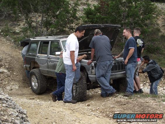 Several guys looking at a lifted Jeep Cherokee XJ with its hood open, parked on a dirt trail
