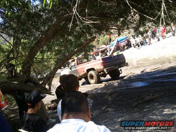 Red and blue pickup truck with large tires on a muddy trail, rear view