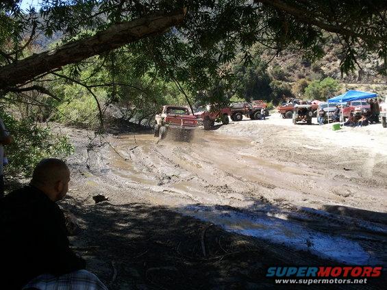 Several off-road trucks driving through a muddy track with spectators watching