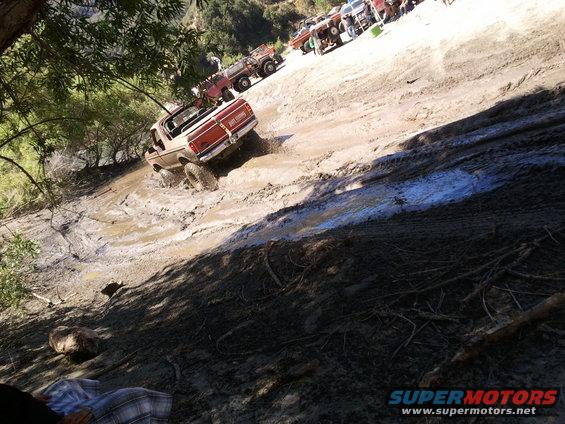 Trucks navigating a muddy incline with trees on the side. One truck is in the foreground, others are further up the slope