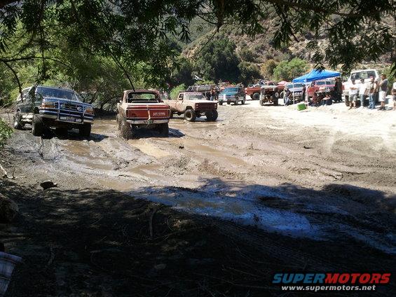 Red pickup truck with lifted suspension driving through mud, front black SUV on incline, other vehicles and people in background