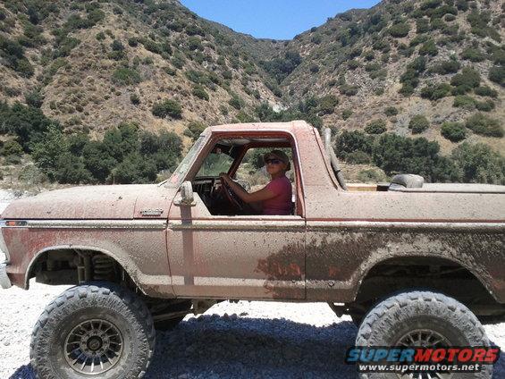 Red 1978 Ford Bronco covered in dirt, side view, with a woman driving