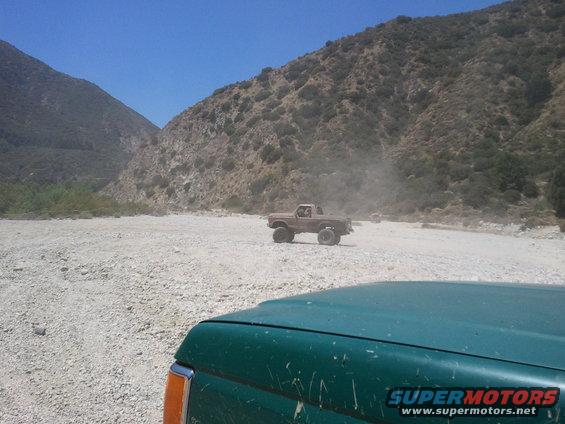 Two trucks driving off-road in a dry riverbed with mountains in the background. One truck is in the foreground, partially visible