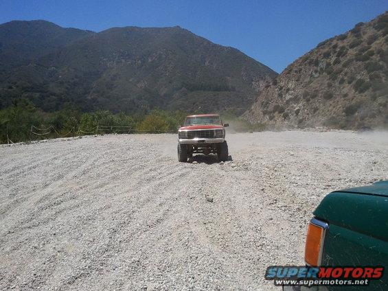 red pickup truck driving on gravel road with mountains behind it, green truck in foreground