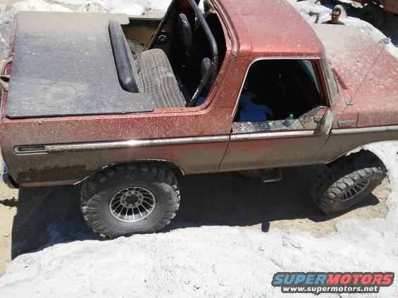 Red 1978 Ford Bronco with a soft top and roll cage, covered in dirt, on a rocky terrain