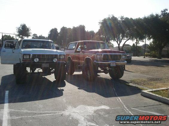 White pickup truck and red Bronco parked side-by-side, both lifted with large tires