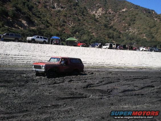 Red 1978 Ford Bronco stuck in mud with other vehicles parked on a ridge in the background