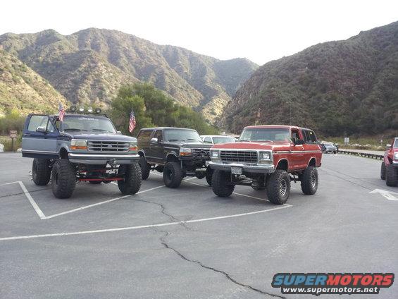Three lifted SUVs parked in a lot with mountains behind them. The red one on the right looks like a 1978 Ford Bronco