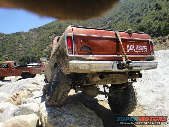 Rear view of a lifted, mud-covered pickup truck with 'ALLEY CUSTOMS Los Angeles, Ca.' decal on tailgate, parked on rocks. Another pickup truck is visible to the left