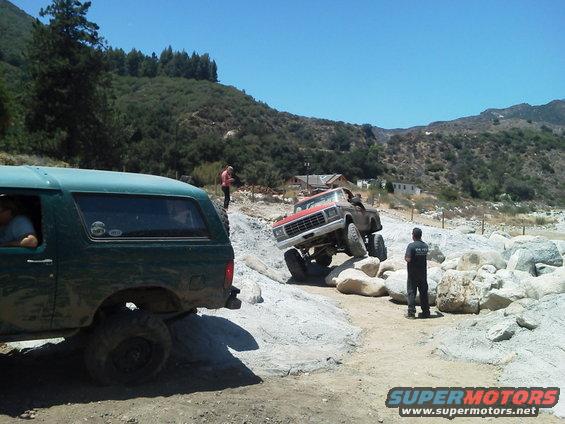 Green Bronco and red-and-brown truck navigating a rocky trail with people watching