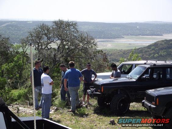 Several Jeep Cherokees parked on a grassy hill with people standing around them; a valley is visible in the background