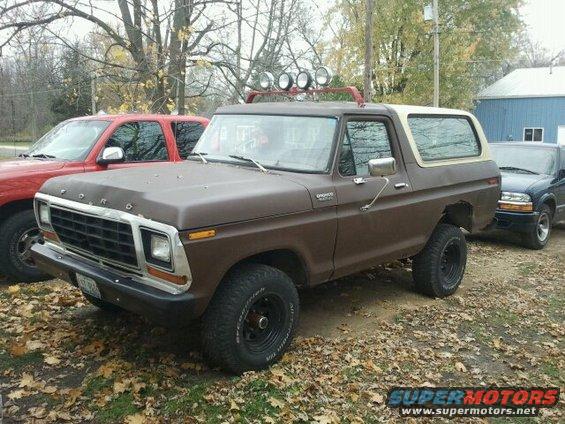 Brown 1978 Ford Bronco with aftermarket lights on roof rack, parked next to red truck and blue SUV