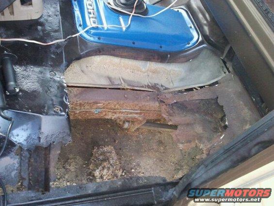 Interior shot of a vehicle's floor with significant rust and damage. Blue panel with 'FORD' visible