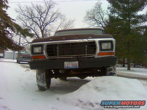 Front view of a brown Ford truck in snow. License plate L17 9406 is visible. Another vehicle is parked behind it